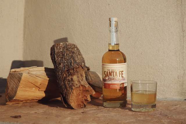 A bottle of Santa Fe whiskey next to a glass half-filled with a golden liquid, set alongside some chopped firewood against a sunlit wall.