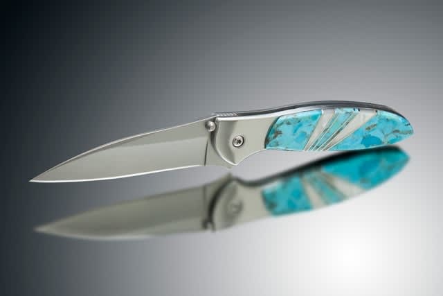 Folding knife with turquoise inlay handle resting on a reflective surface, producing a clear reflection of its blade and handle.