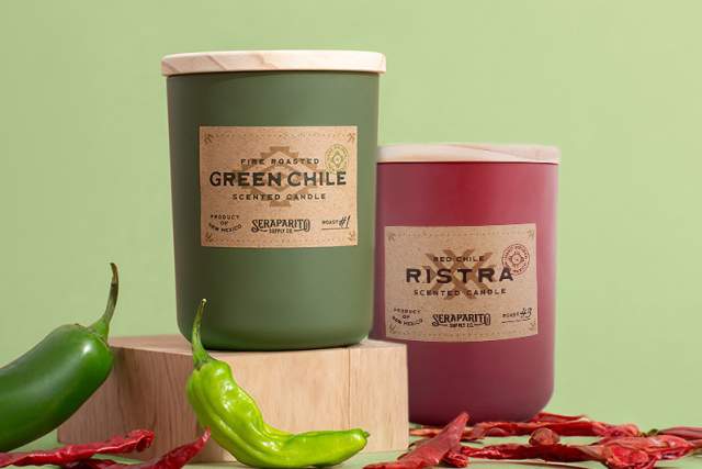 Two scented candles labeled "Green Chile" and "Red Chile Ristra" on a table, with fresh and dried chiles nearby. The background is soft green.