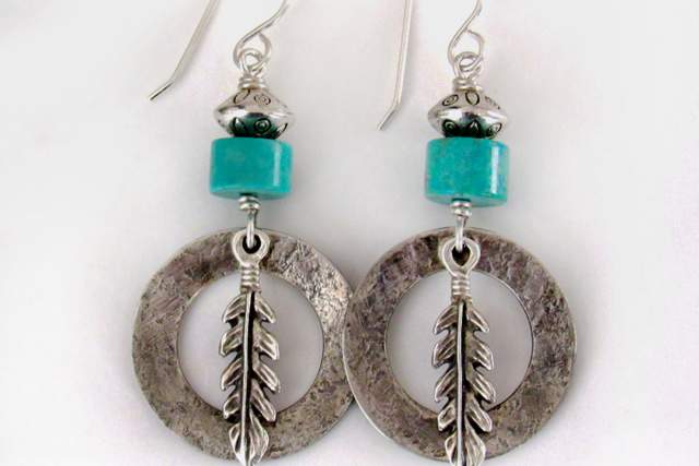 A pair of earrings featuring hammered silver hoops with a feather charm, topped with a turquoise cube and small black bead, on silver hooks.