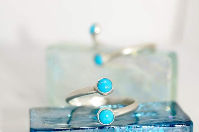 Two twisting sterling silver rings with a turquoise stone, displayed on a matching glass jewelry box under soft lighting.