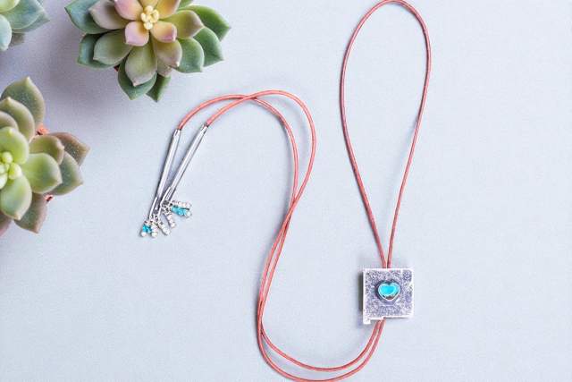 A necklace with a silver pendant shaped like the state of New Mexico featuring a blue heart-shaped gem, displayed on a light background flanked by two succulent plants.