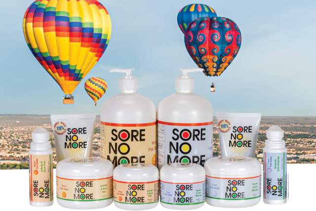 An array of Sore No More natural pain relief products displayed in the foreground, including bottles and jars of varying sizes with labels prominently visible. In the background, colorful hot air balloons float in a clear sky above a landscape.