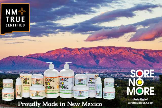 Sore No More product lineup displayed against a scenic landscape in New Mexico, with a mountain range and cloudy sky during sunset. The image includes the "NM True Certified" logo. Text: "Proudly Made in New Mexico" and "Sore No More Pain Relief".