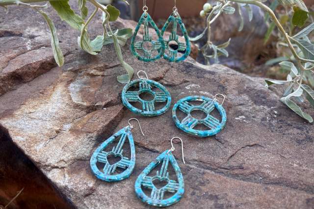Three pairs of crushed turquoise earrings displayed on a stone surface with a green plant in the background. Each pair features a unique design: teardrop shape with a zia symbol and circular insets, round with a cut-out circle, and a larger teardrop design with a heart shape at the center.