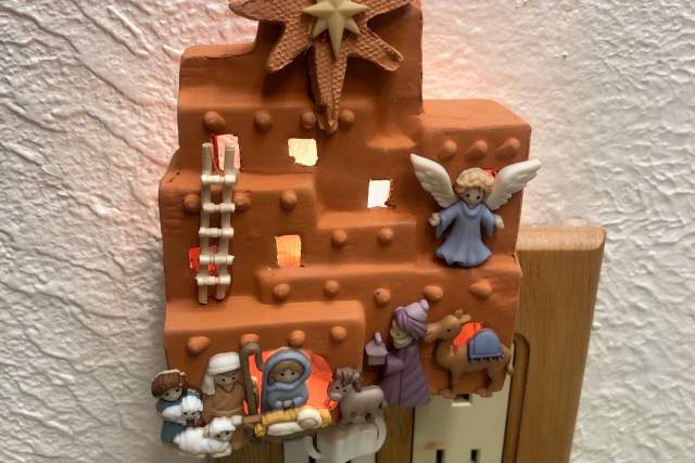Decorative nightlight featuring a nativity scene with various figures, including an angel, placed on an adobe building-shaped structure, illuminated from behind while plugged into a wall outlet.