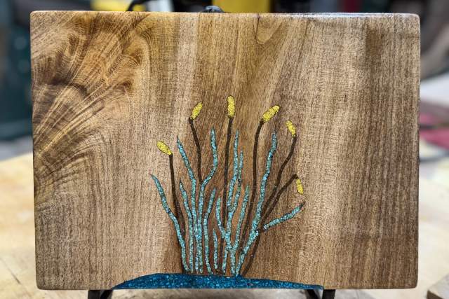 Handcrafted wooden board featuring an artistic inlay of a blue bush and yellow leaves, displayed on a table in a workshop.