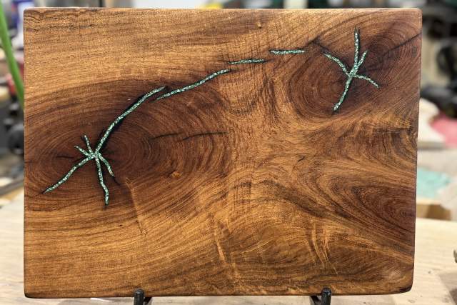Wooden board with a turquoise resin inlay designed to look like a winding branch with leaves, displayed on a stand in a workshop.