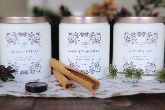 Three scented candles labeled "Holiday Cottage," "Naughty and Nice," and "Winter's Tale" by Storyteller Candles on a table, decorated with pine cones, cinnamon sticks, and holiday greenery.