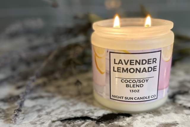 A lit candle labeled "Lavender Lemonade, Coco/Soy Blend, 13 oz" from Night Sun Candle Co. is on a marble surface with blurred lavender sprigs in the background.