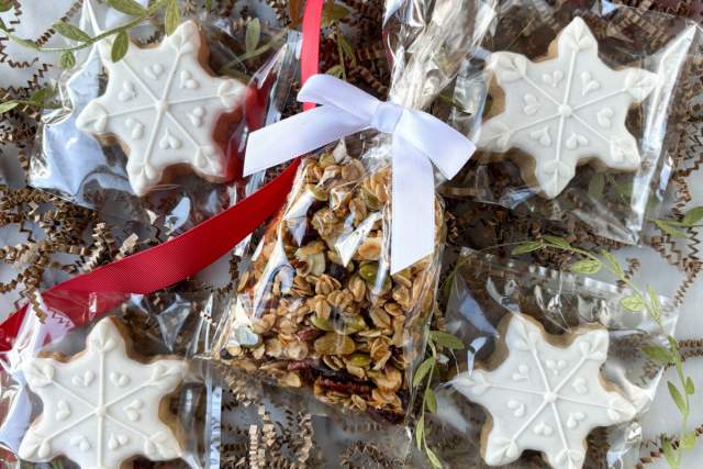 Holiday treats packaged as gifts, featuring snowflake-shaped cookies with white icing and a bag of granola with nuts and seeds, tied with red and white ribbons.