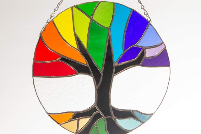 Stained glass artwork featuring a colorful tree with multicolored leaves set against a clear background, suspended by a chain.