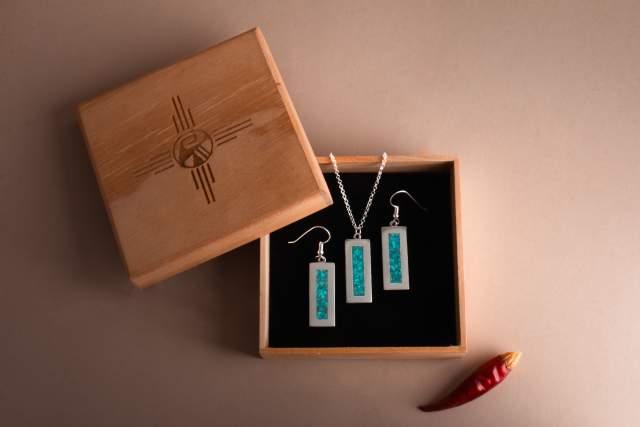 A pair of turquoise and silver earrings displayed in an open wooden box with the Zia sun symbol on the lid, placed next to a red chili pepper on a beige background.