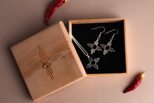 A pair of Zia sun symbol earrings displayed in an open wooden box, with two red chili peppers beside it on a beige background.