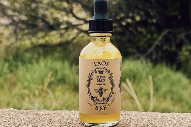 A bottle of Taos Bee Mesa Herbal Beard Balm oil on a stone surface with greenery and purple flowers in the background.
