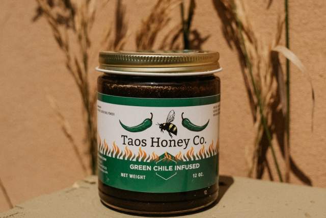 A jar of Taos Honey Co. green chile infused honey, set against an adobe backdrop with dried plants in the background.