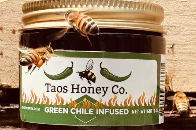 A jar labeled "Taos Honey Co. Green Chile Infused" with illustrations of green chiles and flames. Several bees are flying around the jar, which is sitting on a wooden surface.