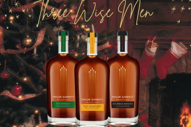 Three bottles of Taylor Garrett whiskey (Rye, Old Fashioned, Bourbon) displayed in front of a festive Christmas background with twinkling lights, a tree, and a cozy fireplace.