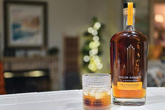 A bottle of Taylor Garrett Old Fashioned whiskey sits on a countertop next to a textured glass filled with a drink and ice cubes. The background is softly blurred with warm indoor lighting.