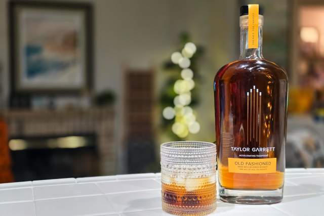 A bottle of Taylor Garrett Old Fashioned whiskey next to a glass half-filled with the beverage, set on a table with a softly blurred background featuring indoor lighting and holiday decorations.