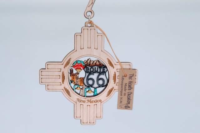 A wooden ornament depicting the Zia symbol with colorful illustrations including mountains and the words "Route 66 New Mexico." A small tag is attached.