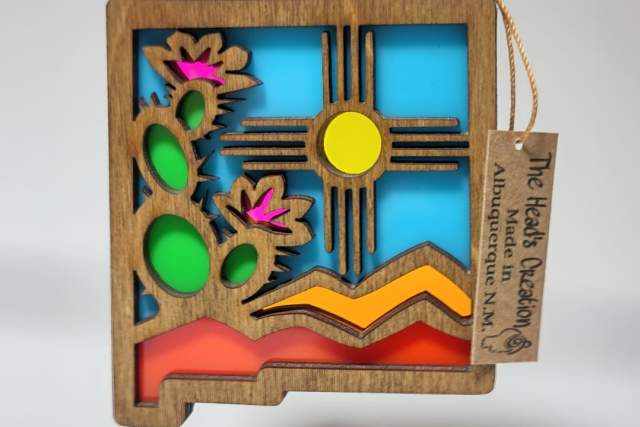 A colorful, layered wooden ornament featuring the Zia sun symbol, with elements in shades of blue, green, yellow, pink, and orange. A tag attached reads "The Hands of Creation ABQ NM.