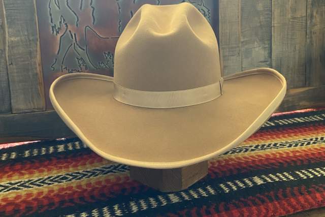 A cowboy hat rests on a wooden beam in front of a colorful woven textile and a framed artwork featuring a stylized face.