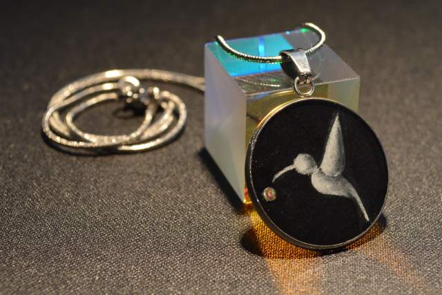 A silver necklace with a pendant featuring a hummingbird design is draped over a small, translucent cube.