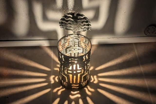 A decorative candle holder casts intricate, shadowy patterns on a light-colored wall, with beams radiating outward. The patterns resemble a complex, geometric design.