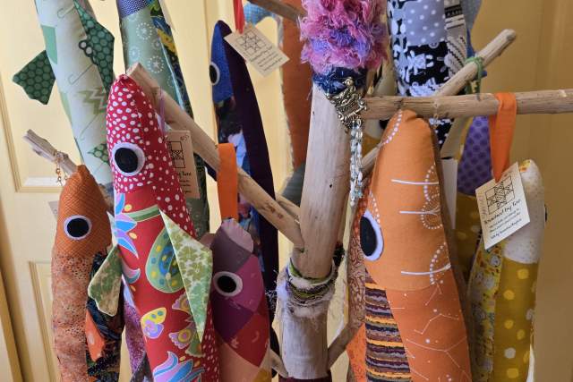 A group of colorful, patterned stuffed fish toys are hanging from wooden branches. The display is indoors, in front of a beige door.