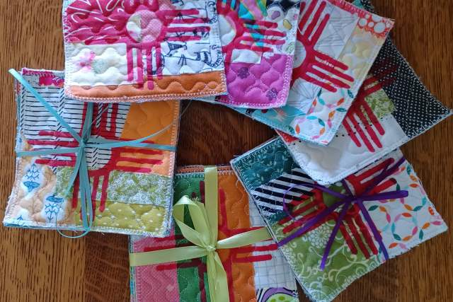 A collection of colorful, handmade cloth coasters displayed on a wooden table, each uniquely patterned and some tied with ribbons.