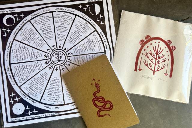 A photo featuring a circular witch's almanac chart, a notebook with a serpent design on its cover, and a napkin with a tree printed on it, all laid out on a dark surface.
