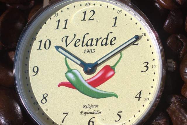 A wristwatch with a brown leather strap is surrounded by coffee beans. The watch face features the brand name "Velarde," the year "1903," and a design with two chili peppers, one green and one red. "Relojeros Espléndidos" and "New Mexico" are also visible on the dial.