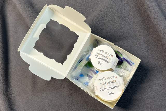 An open cardboard package on a gray fabric surface containing two eco-friendly products: a "Well Worn Naturals Shampoo Bar" and a "Well Worn Naturals Conditioner Bar".