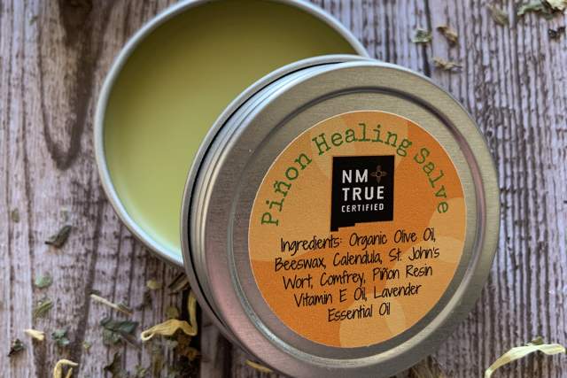 An open tin of Piñon Healing Salve on a wooden surface surrounded by dried herbs. The label lists organic ingredients like olive oil, beeswax, and lavender essential oil.