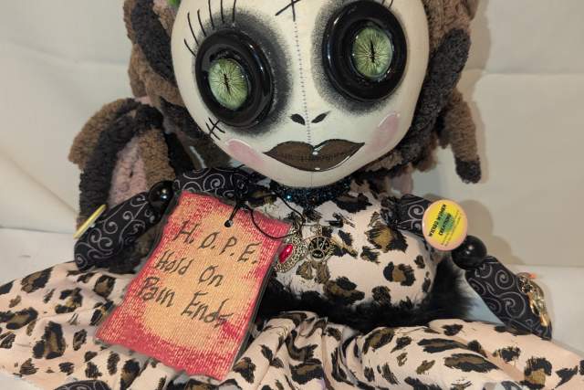 Plush doll with stitched facial features, multicolored yarn hair, and animal print outfit, sitting against a white background. The doll holds a tag labeled 'HOPE Hold on Pain Ends.'