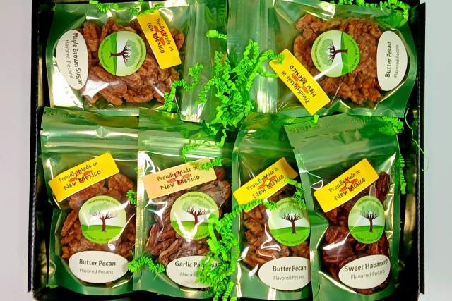 Display of various flavored pecans in clear packaging, including Butter Pecan, Garlic Pecan, and Sweet Habanero, each labeled with bright stickers and arranged neatly in a dark tray with decorative green accents.
