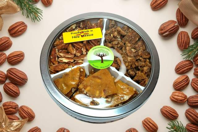 A round tin filled with assorted traditional New Mexican treats including biscochitos, pine nuts, and pecan brittle, labeled as products made in New Mexico. The tin is surrounded by pecans and festive decorations.