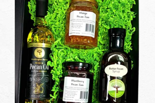 Gift box containing bottles of Pecan Oil, Blackberry Puree, Honey Ginger Sauce, and Butter Pecan Syrup, set in green decorative packaging with a festive Christmas border featuring ornaments and pine branches.