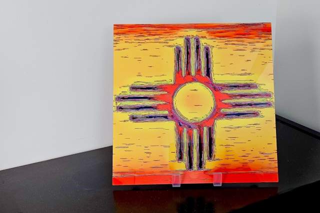 A painting of the Zia Sun symbol, displayed on a stand, featuring a yellow circle center with rays radiating outward set against a vibrant yellow and orange background.