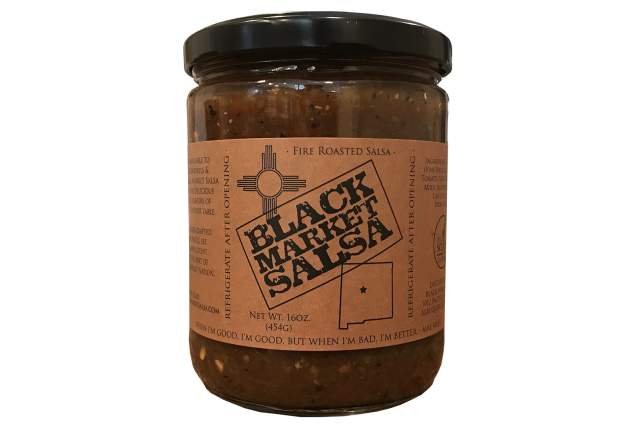 A jar filled with dark red salsa, with a black top. The red label, with a Zia Symbol and the state of New Mexico, reads Black Market Salsa, Fire Roasted Salsa "When I'm good, I'm good, When I'm Bad, I'm even better" - Mae West