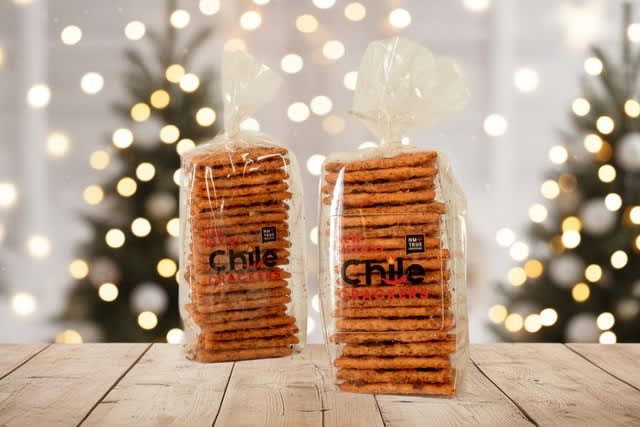 Two packages of Chile Crackers displayed on a wooden table, with a blurred background featuring twinkling lights.