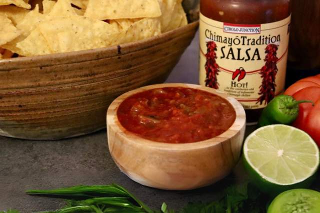 fresh vegetables, fruits and peppers sit before a wooden bowl filled with red salsa next to a wooden bowl of chips. A jar of salsa with a red cap and tan label sits behind them. The label reads "Chimayo Tradition Salsa, HOT!"