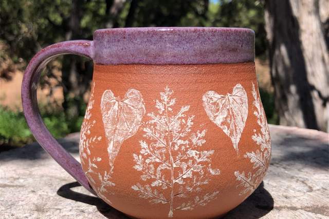 A ceramic mug with a rustic design, featuring botanical prints and heart shapes, set against a natural backdrop with trees.