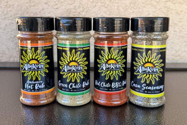 4 clear containers with black, yellow and green or red labels sit on a black countertop with a white spackled background. Label reads "AlbuKirky Seasonings Green/Red Chile Rub, Hot BBQ Rub and Casa Seasoning" respectively