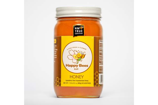 A mason jar filled with golden honey. Stamped with the New Mexico True Certified black logo in the shape of New Mexico. The yellow and brown label with a honey bee hugging a flower reads Happy Bees LLC, a kiss from a flower. Honey, Ingredients: Raw Unpastrurized Honey