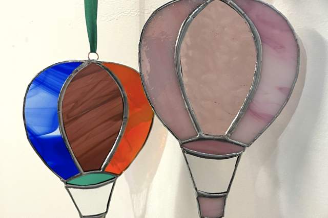 two multicolored stained glass balloons hang from pink and green strings in front of a white background