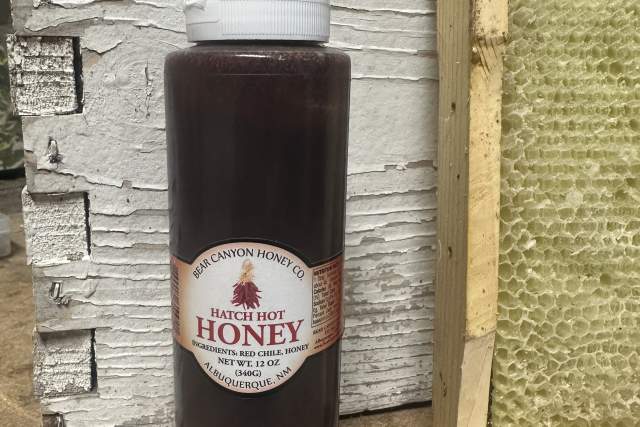 a clear plastic bottle with a squeeze top sits next to a wooden wall and a framed bee honeycomb. The label reads "Bear Canyon Honey Co. Hatch Hot Honey"