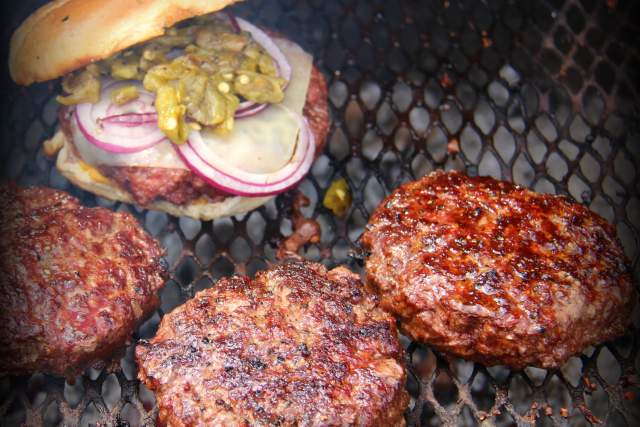 Four hamburgers sit on a grill grate, on sitting on a bun with cheese, green chile and red onion.