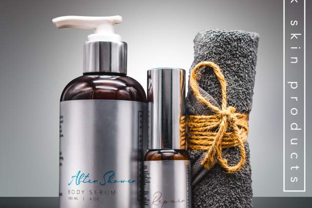 Alt Text: "K+K Skin Products Men's Gift Set featuring an amber bottle of body serum, a metallic bottle of after shave, and a dark gray towel tied with twine. All items are set against a gradient gray background.
